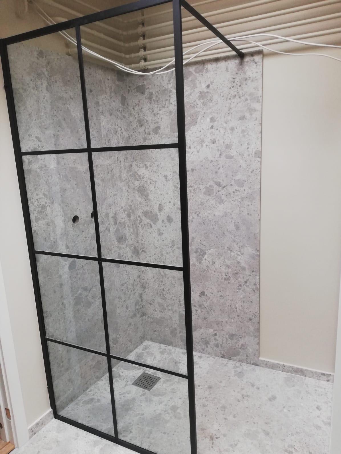 Nowoczesna kabina prysznicowa z czarną ramą i szklanymi panelami, wykończona płytkami w odcieniach szarości z wyraźnym wzorem marmuru, widoczny odpływ liniowy w podłodze.