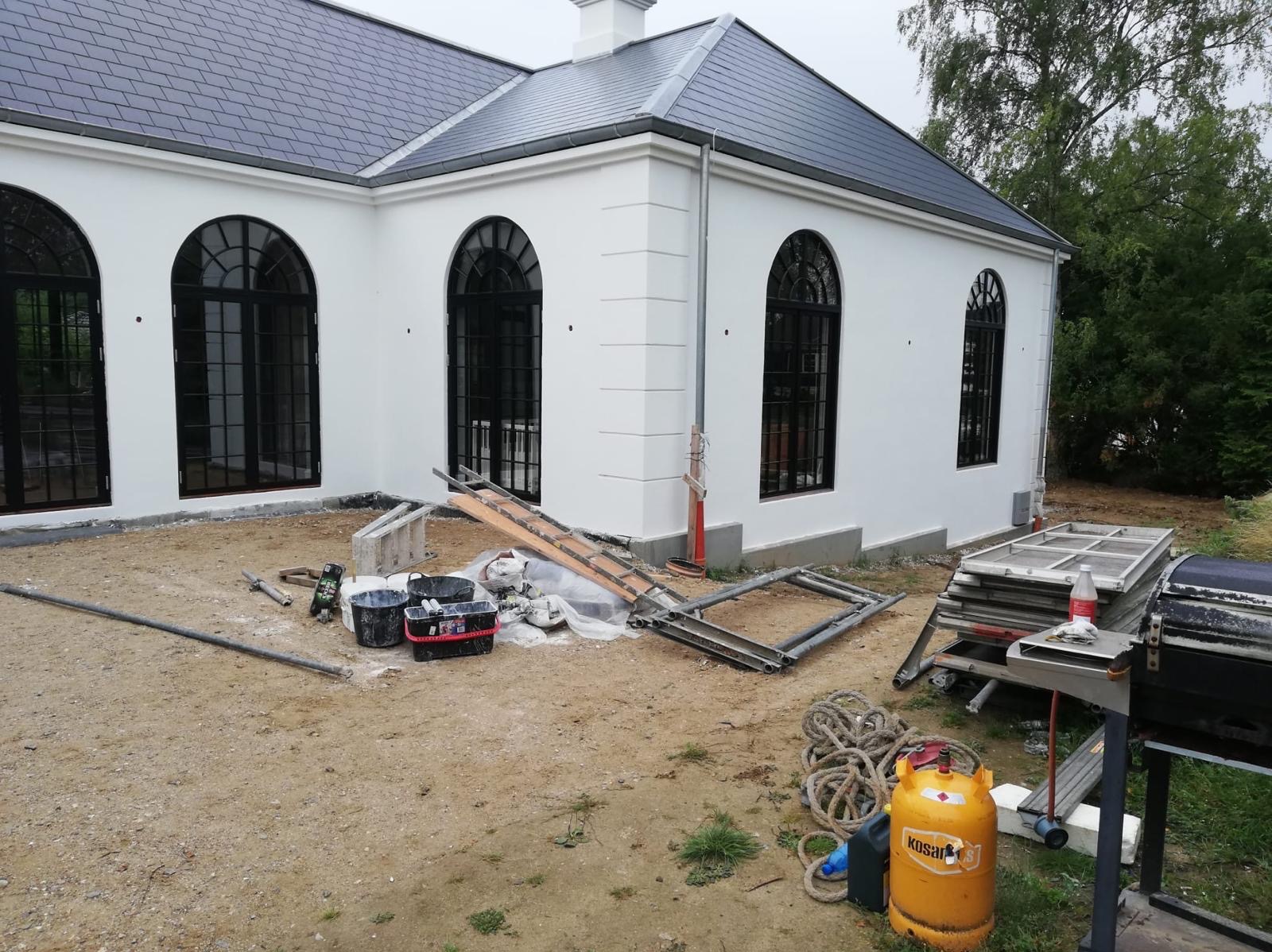 Elewacja budynku z czarnymi, łukowatymi oknami po malowaniu, z widocznymi narzędziami i materiałami budowlanymi na ziemi, butla gazowa Kosan na pierwszym planie.