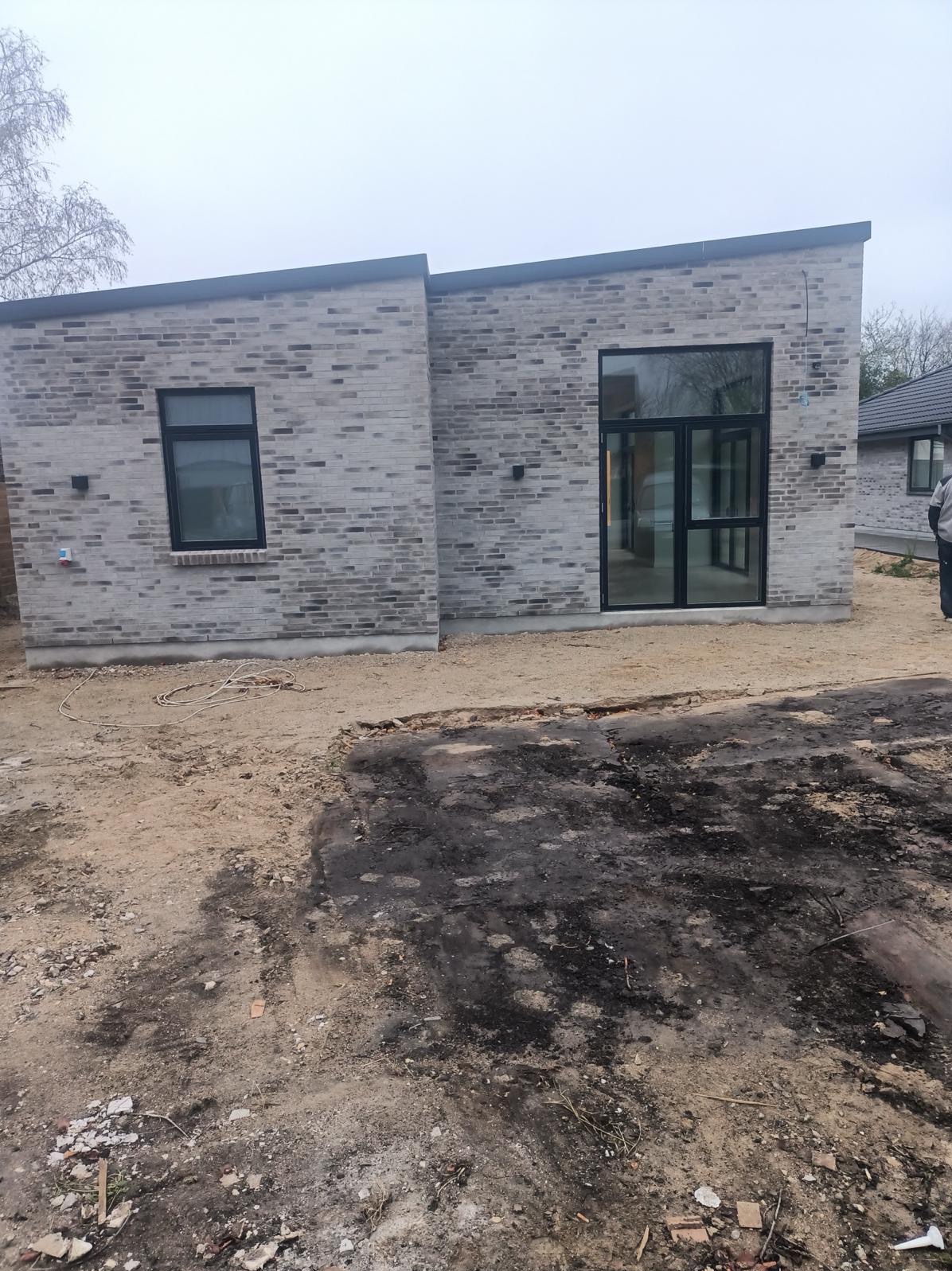 Nowoczesny dom z szarej cegły z czarnymi ramami okiennymi i drzwiowymi, teren wokół w trakcie prac ziemnych.