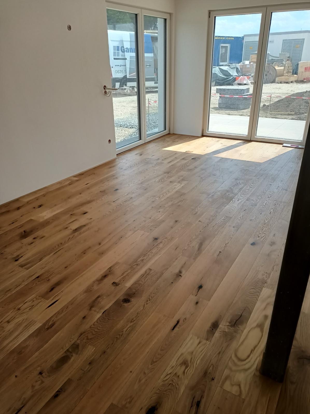 Nowo położona, jasna, drewniana podłoga z widocznymi sękami i naturalnym usłojeniem w pustym pomieszczeniu z dużymi oknami balkonowymi, przez które widać teren budowy.