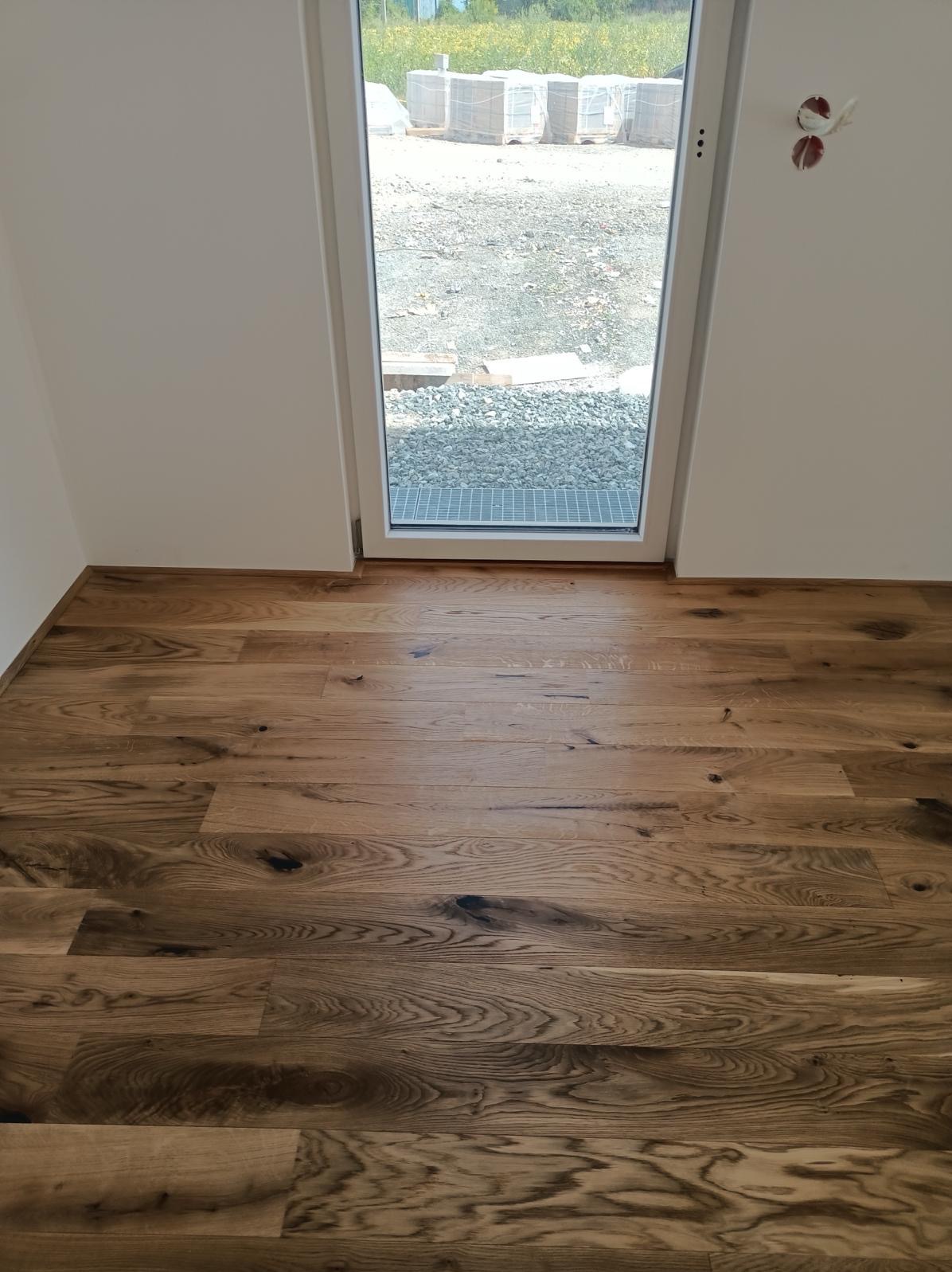 Ułożona drewniana podłoga z widocznymi słojami i sękami, z fragmentem okna wychodzącego na plac budowy z paletami cegieł.