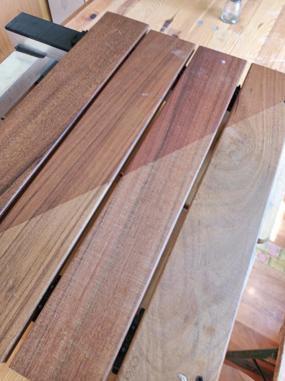 Cztery drewniane deski połączone czarnymi metalowymi elementami, z widoczną różnicą w odcieniach brązu po malowaniu lub lakierowaniu, ułożone na drewnianym stole, z butelką w tle.