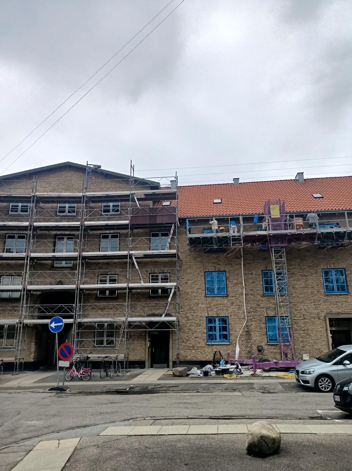 Dwa budynki z cegły, jeden z rusztowaniem, drugi z podnośnikiem, okna zabezpieczone folią, prace na wysokościach w toku.