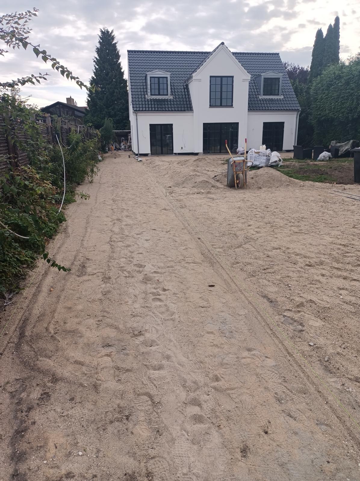 Nowoczesny dom jednorodzinny z czarnymi oknami i drzwiami, widok z zewnątrz na przygotowywany teren pod ogród, z widoczną taczką, workami i śladami opon w piasku.