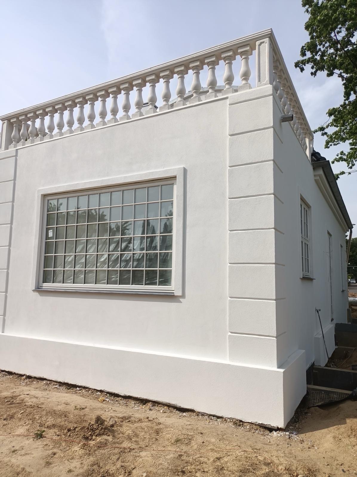 Biała fasada budynku z balustradą na dachu, okno z drobnymi podziałami, widoczne ślady niedawnego szpachlowania i malowania.