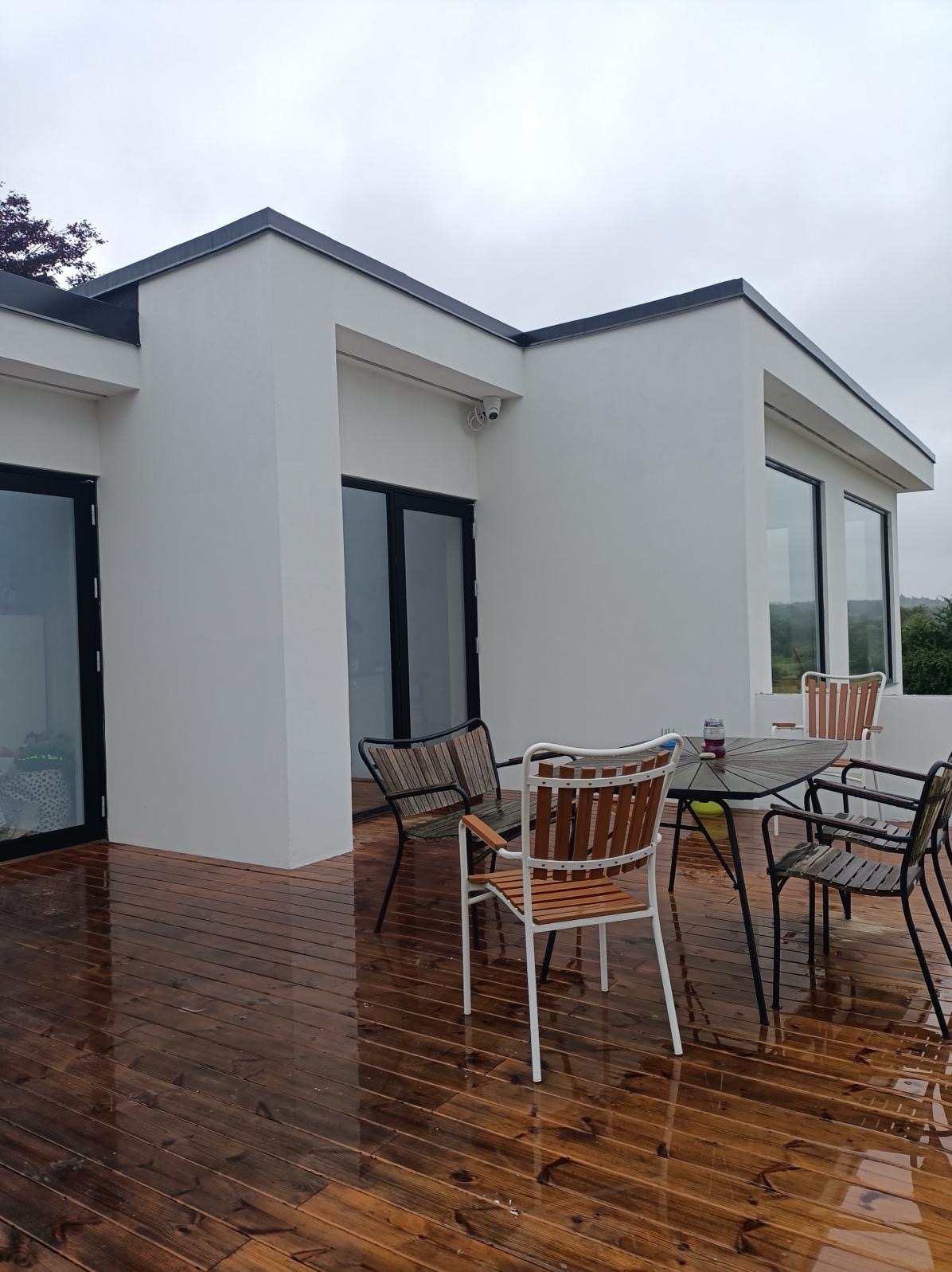 Nowoczesny dom z białymi ścianami i drewnianym tarasem po deszczu, widoczne krzesła i stół ogrodowy.