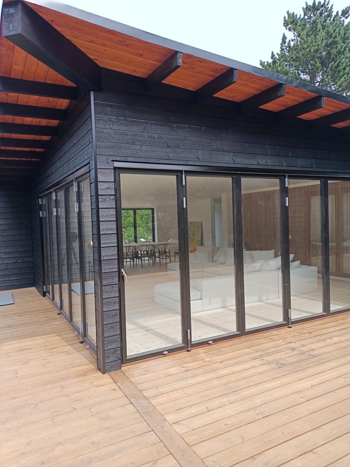 Czarny, drewniany dom z rozsuwanymi, szklanymi drzwiami wychodzącymi na drewniany taras; widok na jasne wnętrze z sofą i stołem w tle.