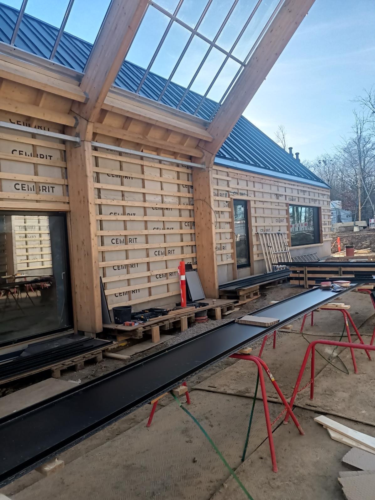 Budynek w trakcie budowy: widoczne drewniane elementy konstrukcyjne, płyty izolacyjne z nadrukiem, czarne profile metalowe na kozłach, okna i przeszklony dach.