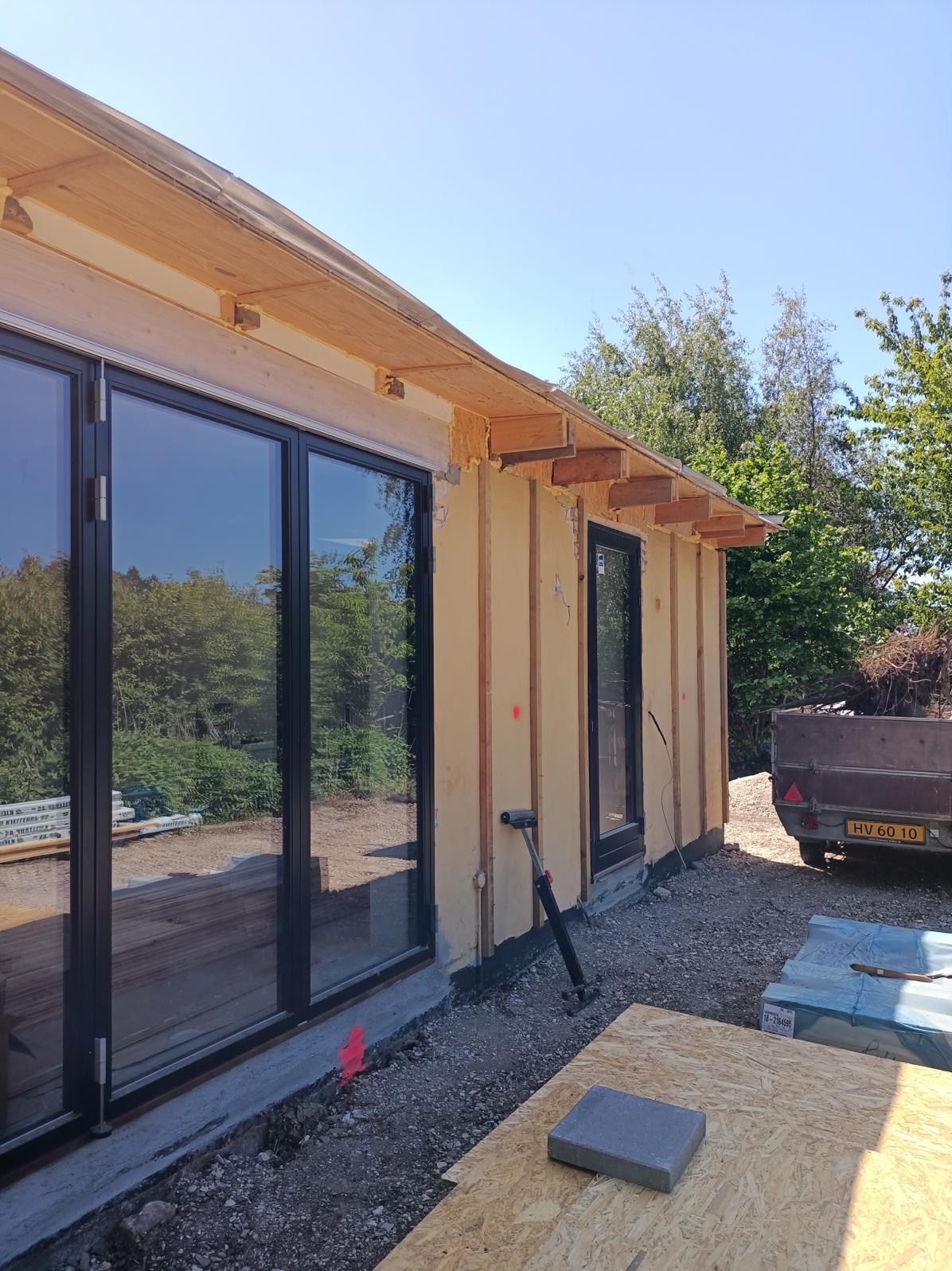 Budynek w trakcie budowy, widoczne duże okna z czarnymi ramami, ściany w surowym stanie z widocznym ociepleniem i drewnianą konstrukcją, na zewnątrz przyczepa z zielenią i materiały budowlane.