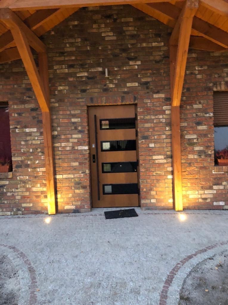 Elewacja domu z cegły klinkierowej z drewnianą konstrukcją zadaszenia i nowoczesnymi drzwiami wejściowymi z przeszkleniami, oświetlona punktowymi lampami gruntowymi.