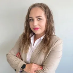 Portret kobiety w beżowym żakiecie i białej koszuli, z założonymi rękami, na jasnoszarym tle.