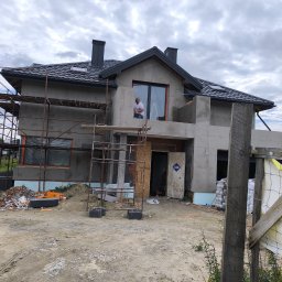 RafBud - Dom w trakcie budowy z rusztowaniem. Widoczny fragment elewacji z oknem,  nieotynkowana ściana,  mężczyzna w oknie i materiały budowlane na ziemi.