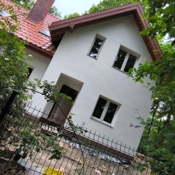 RafBud - Biała fasada domu z czerwonym dachem, widoczna zza metalowego ogrodzenia, otoczona zielenią. Ujęcie z dołu, podkreślające odnowioną elewację i detale architektoniczne.