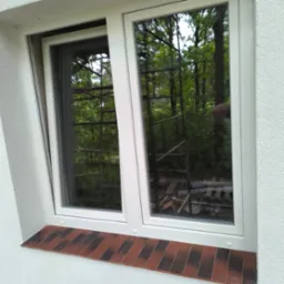 Białe okno PCV z brązowym parapetem z cegły klinkierowej, widoczne rusztowanie i zieleń za oknem. Okno uchylone, ściana z wyraźną fakturą.