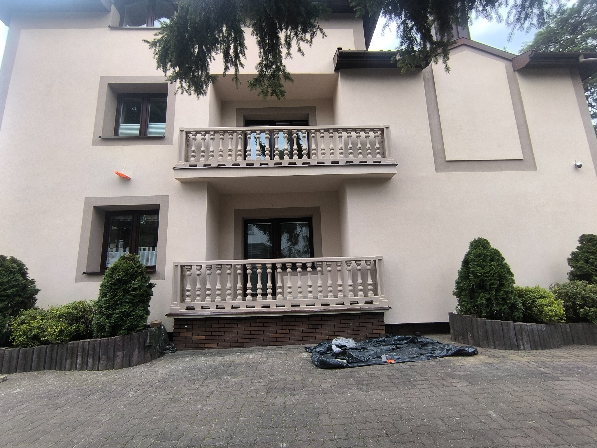 Elewacja domu z dwoma balkonami z balustradami. Jasna kolorystyka, ozdobne obramowania okien, krzewy przed budynkiem. Na chodniku leży czarna folia.
