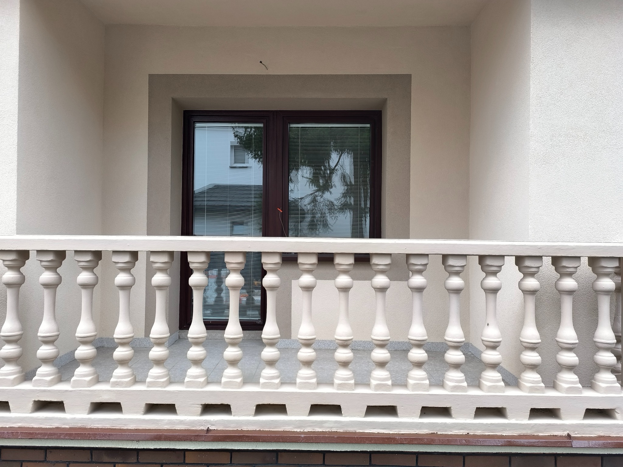 Elegancki balkon z beżową balustradą tralkową i oknem z brązową ramą, widok z dołu, kremowa elewacja budynku w tle.