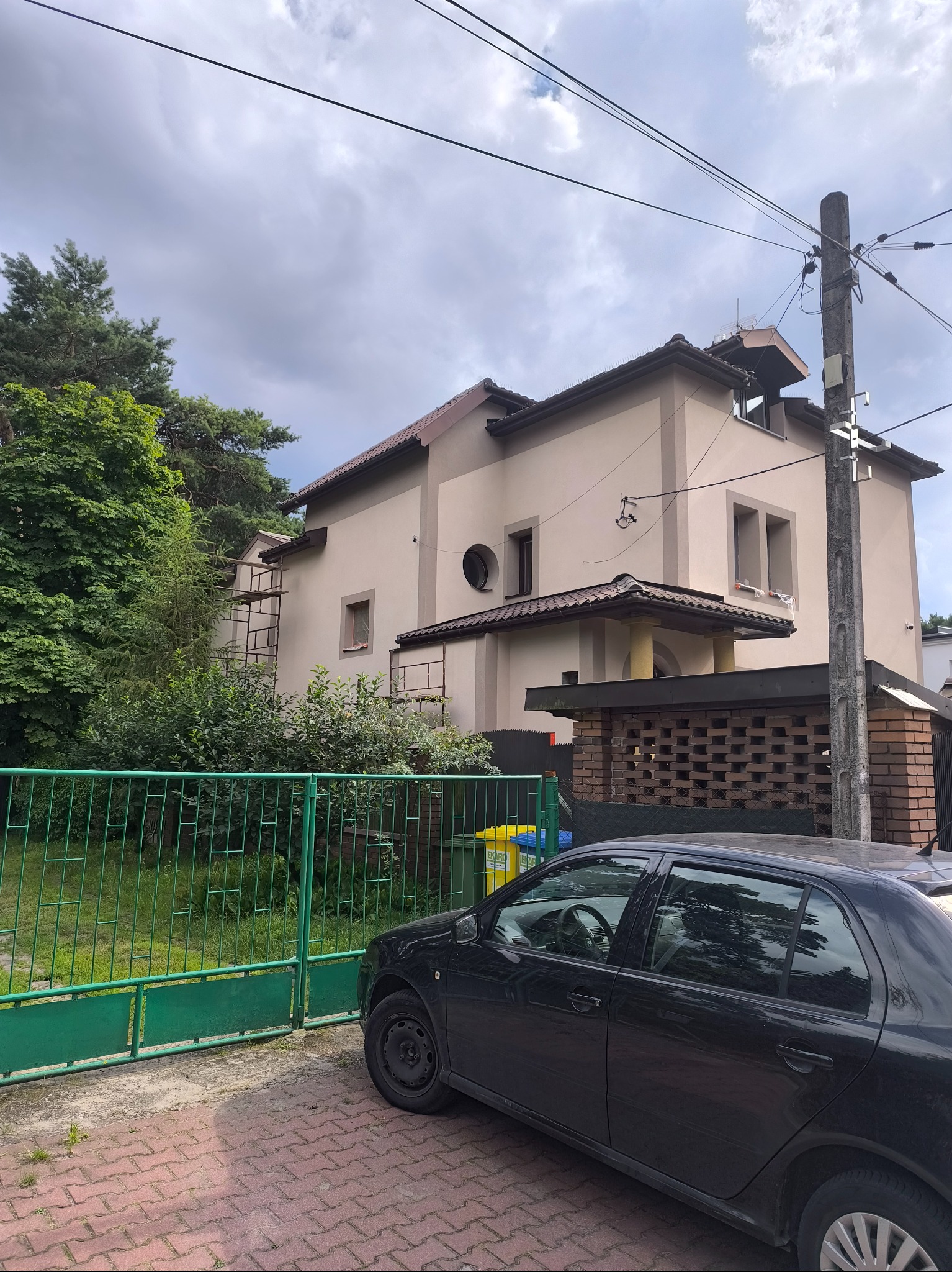 Dwupiętrowy dom z beżową elewacją, widoczne rusztowania przy ścianie. Na pierwszym planie zielony płot i zaparkowany czarny samochód osobowy. Pochmurne niebo w tle.