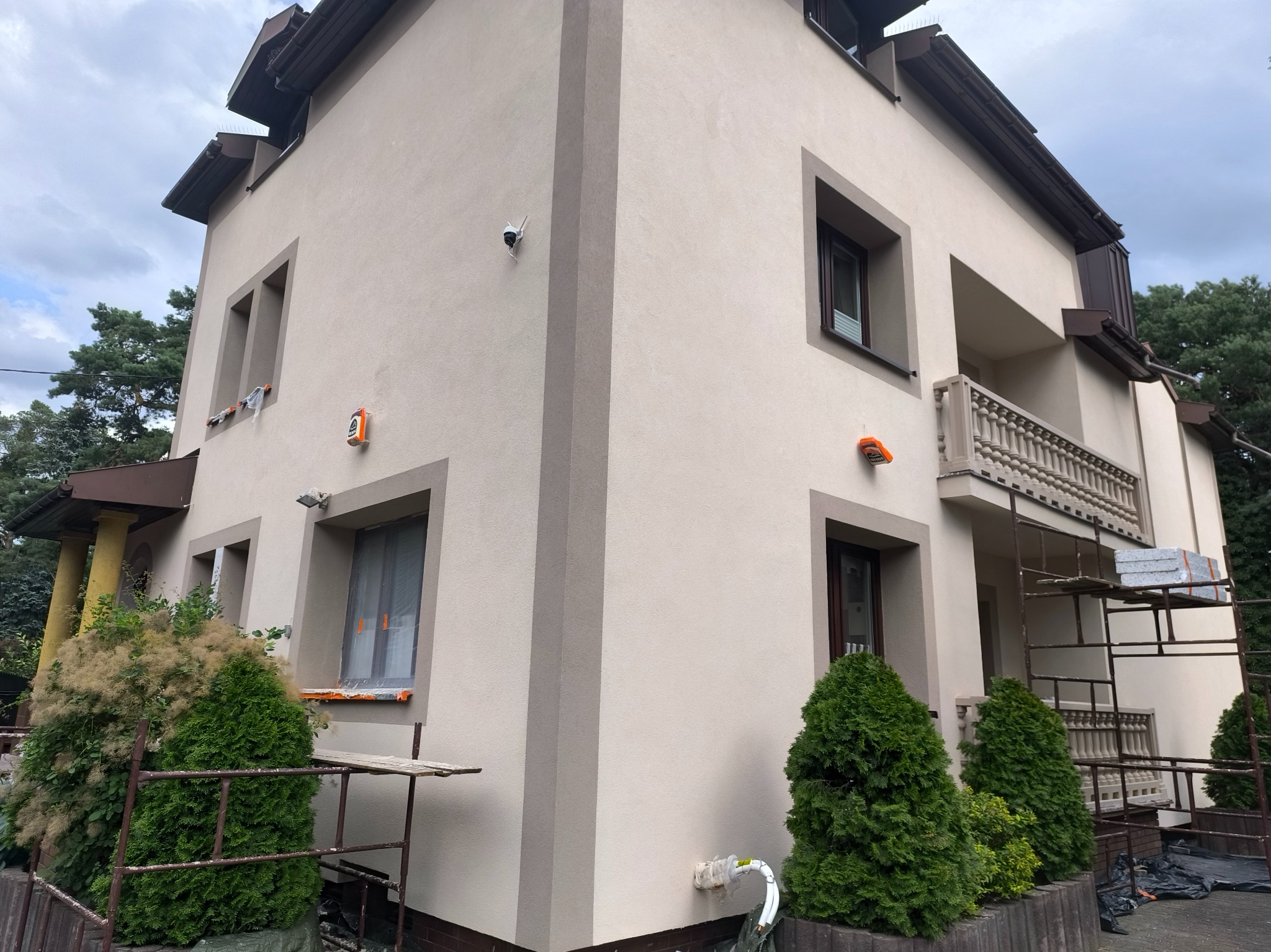 Dwukondygnacyjny dom po renowacji elewacji w odcieniach beżu i brązu, z widocznymi rusztowaniami i zabezpieczonymi oknami, otoczony zielenią.