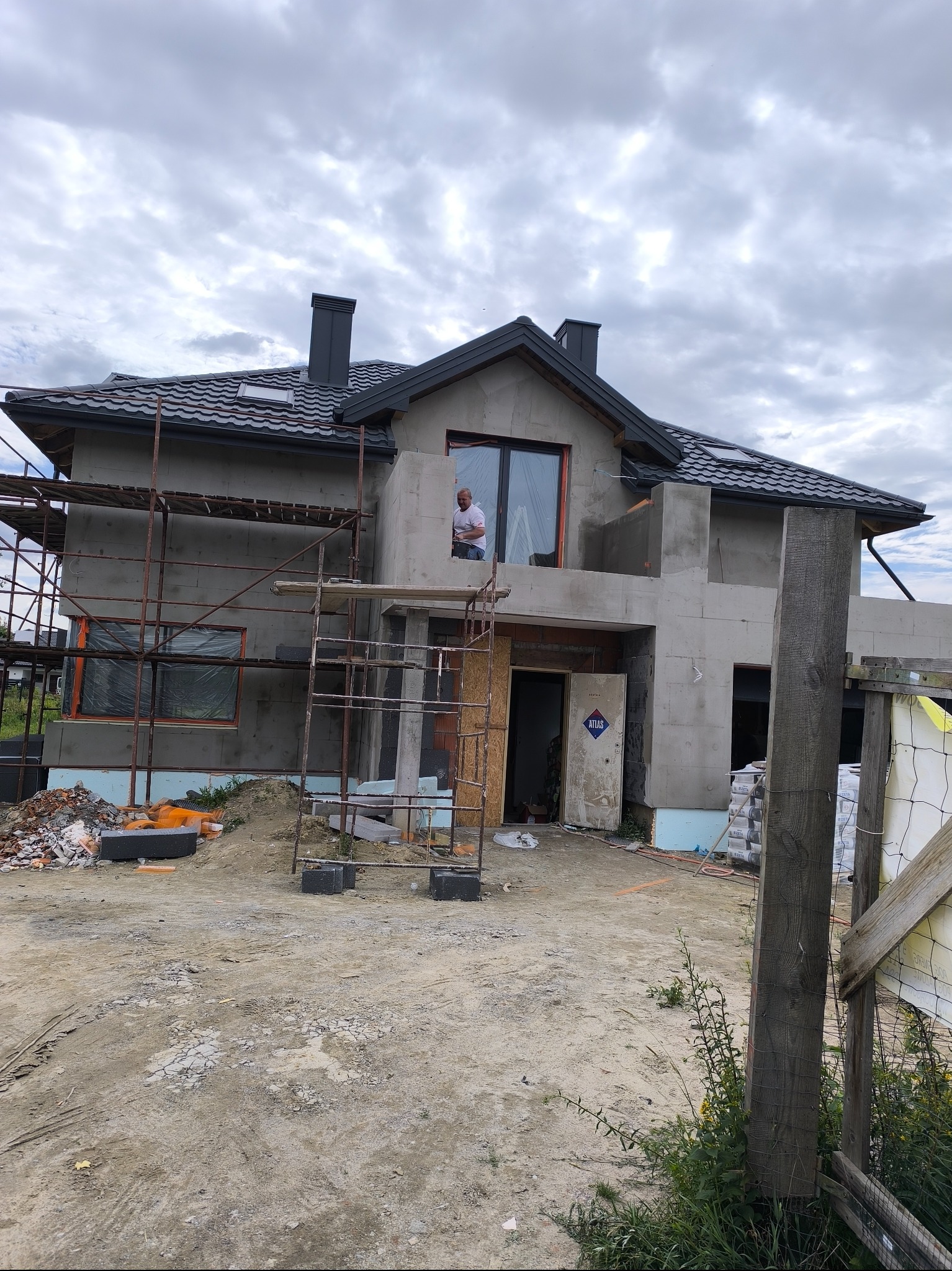 Dom w trakcie budowy z rusztowaniem. Widoczny fragment elewacji z oknem,  nieotynkowana ściana,  mężczyzna w oknie i materiały budowlane na ziemi.
