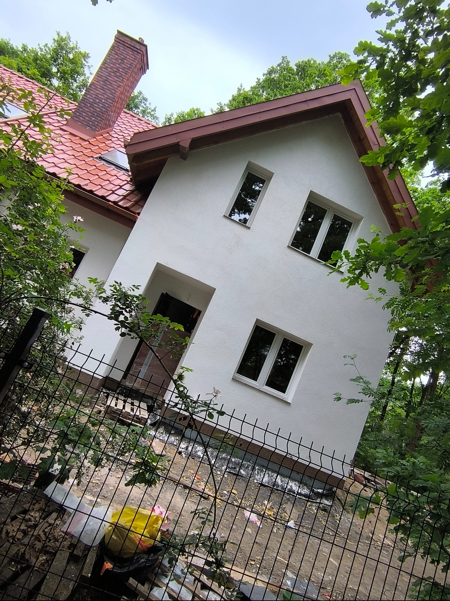 Biała fasada domu z czerwonym dachem, widoczna zza metalowego ogrodzenia, otoczona zielenią. Ujęcie z dołu, podkreślające odnowioną elewację i detale architektoniczne.