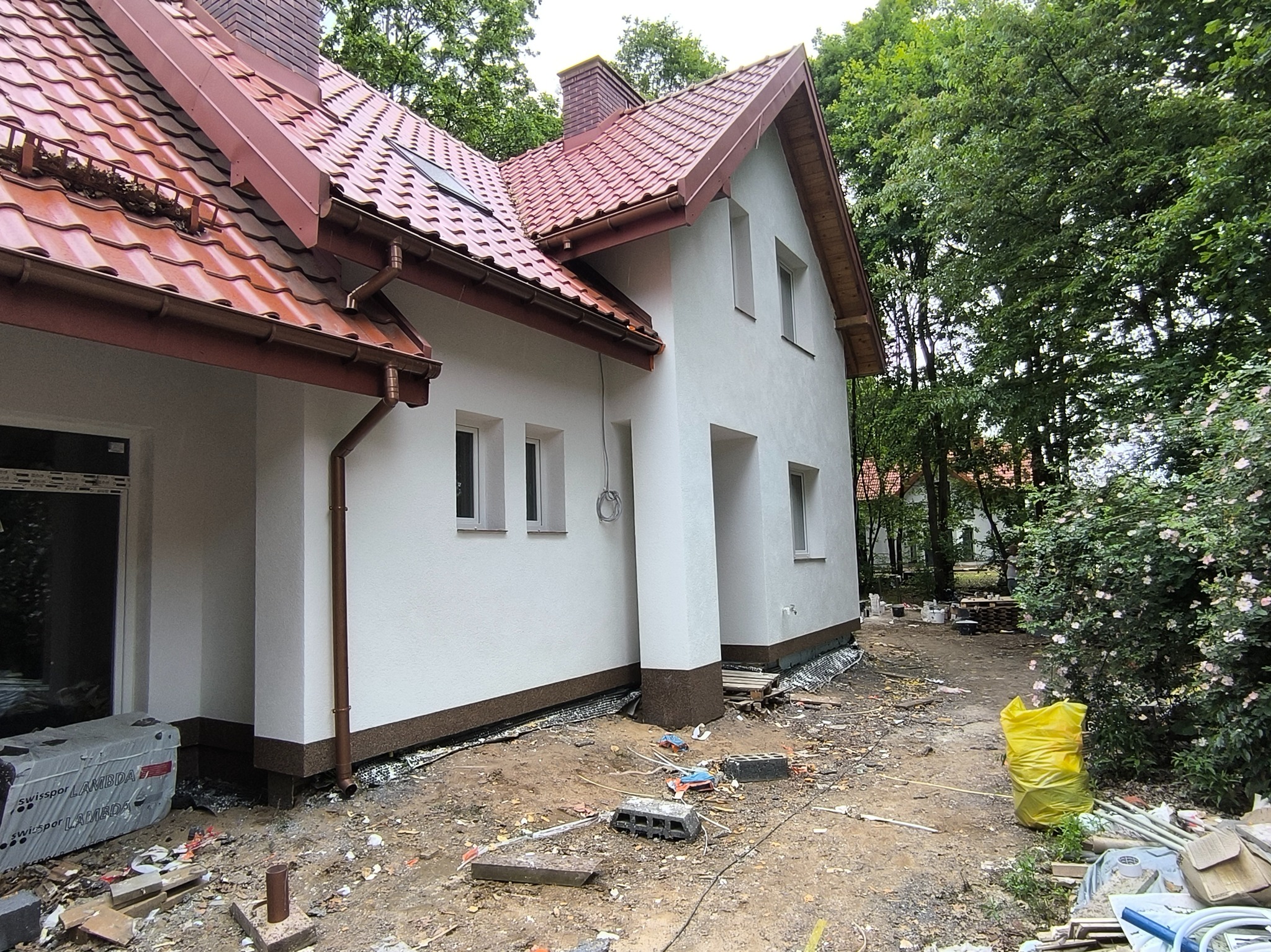 Biały dom z czerwoną dachówką, widok z boku, z elementami wykończenia elewacji i detalami architektonicznymi. Otoczenie w trakcie prac.