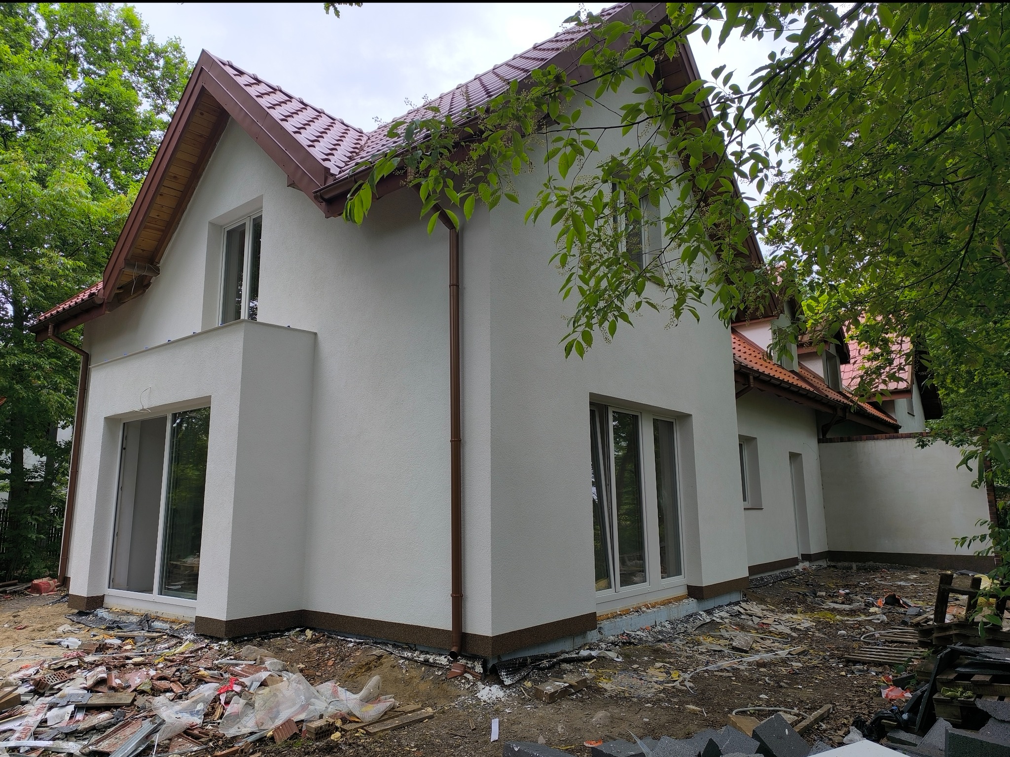 Biały dom jednorodzinny z brązowym dachem i rynnami, otoczony zielenią. Widoczne okna i fragment elewacji. Teren wokół domu w trakcie prac wykończeniowych.
