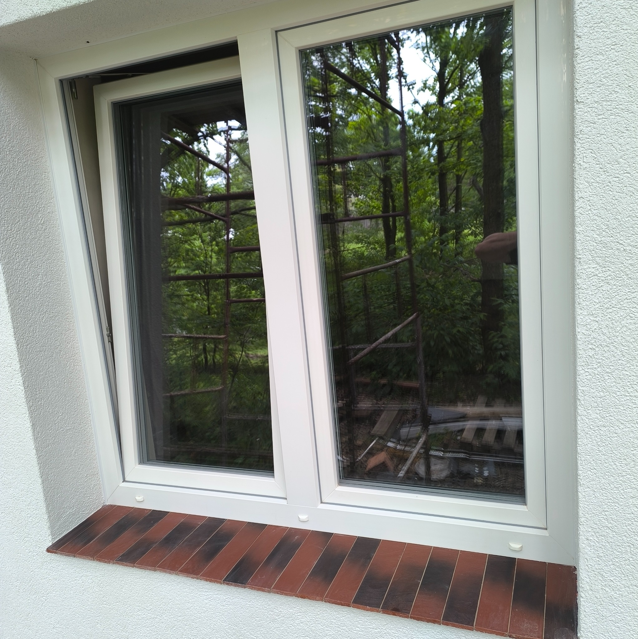 Białe okno PCV z brązowym parapetem z cegły klinkierowej, widoczne rusztowanie i zieleń za oknem. Okno uchylone, ściana z wyraźną fakturą.