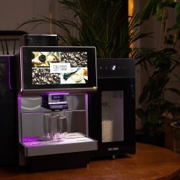 Crazy Fresh - Profesjonalny ekspres do kawy z ekranem dotykowym prezentujący menu, podświetlony na fioletowo, z dwoma szklankami na podstawce, ustawiony na drewnianym stole w otoczeniu roślin doniczkowych.