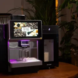 Profesjonalny ekspres do kawy z ekranem dotykowym prezentujący menu, podświetlony na fioletowo, z dwoma szklankami na podstawce, ustawiony na drewnianym stole w otoczeniu roślin doniczkowych.