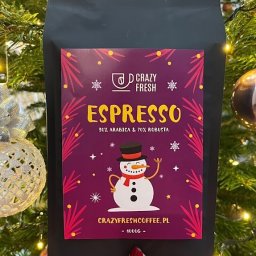 Crazy Fresh - Czarna paczka kawy Espresso 30% Arabica i 70% Robusta marki Crazy Fresh z motywem świątecznym (bałwanek, śnieżynki) trzymana w dłoni na tle choinki z lampkami i bombkami.