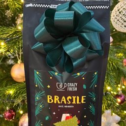 Crazy Fresh - Czarna paczka kawy 'Brasile 100% Arabica' od Crazy Fresh, z zieloną kokardą, na tle choinki z ozdobami, prezentowana w dłoni.