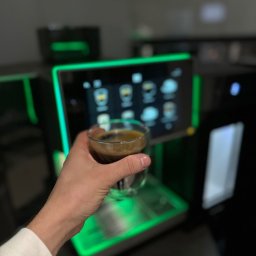 Crazy Fresh - Ręka trzyma szklankę espresso przed podświetlonym na zielono ekspresem do kawy z dotykowym ekranem.