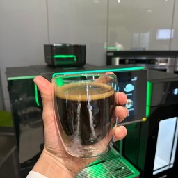 Crazy Fresh - Ręka trzyma szklankę świeżo zaparzonej kawy na tle nowoczesnego ekspresu z podświetleniem LED, widoczny blat kuchenny i białe szafki.