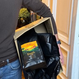 Crazy Fresh - Kurier w brązowym swetrze i jeansach trzyma karton wypełniony szczelnie zapakowanymi czarnymi torbami kawy 'Brasile 100% Arabica' marki Crazy Fresh.