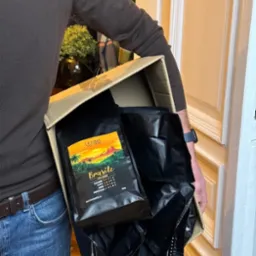 Kurier w brązowym swetrze i jeansach trzyma karton wypełniony szczelnie zapakowanymi czarnymi torbami kawy 'Brasile 100% Arabica' marki Crazy Fresh.