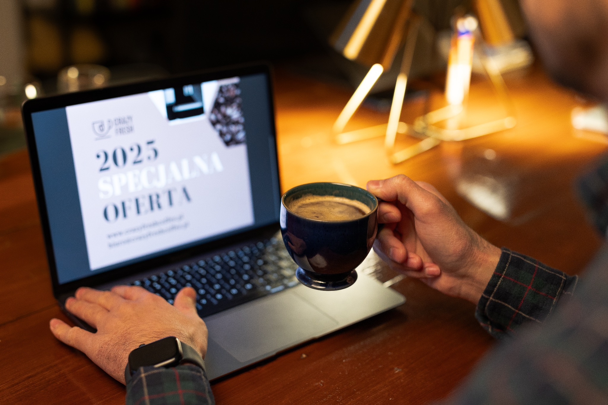 Mężczyzna w koszuli w kratę trzyma filiżankę kawy nad laptopem wyświetlającym ofertę na rok 2025, na drewnianym biurku, w tle lampa.