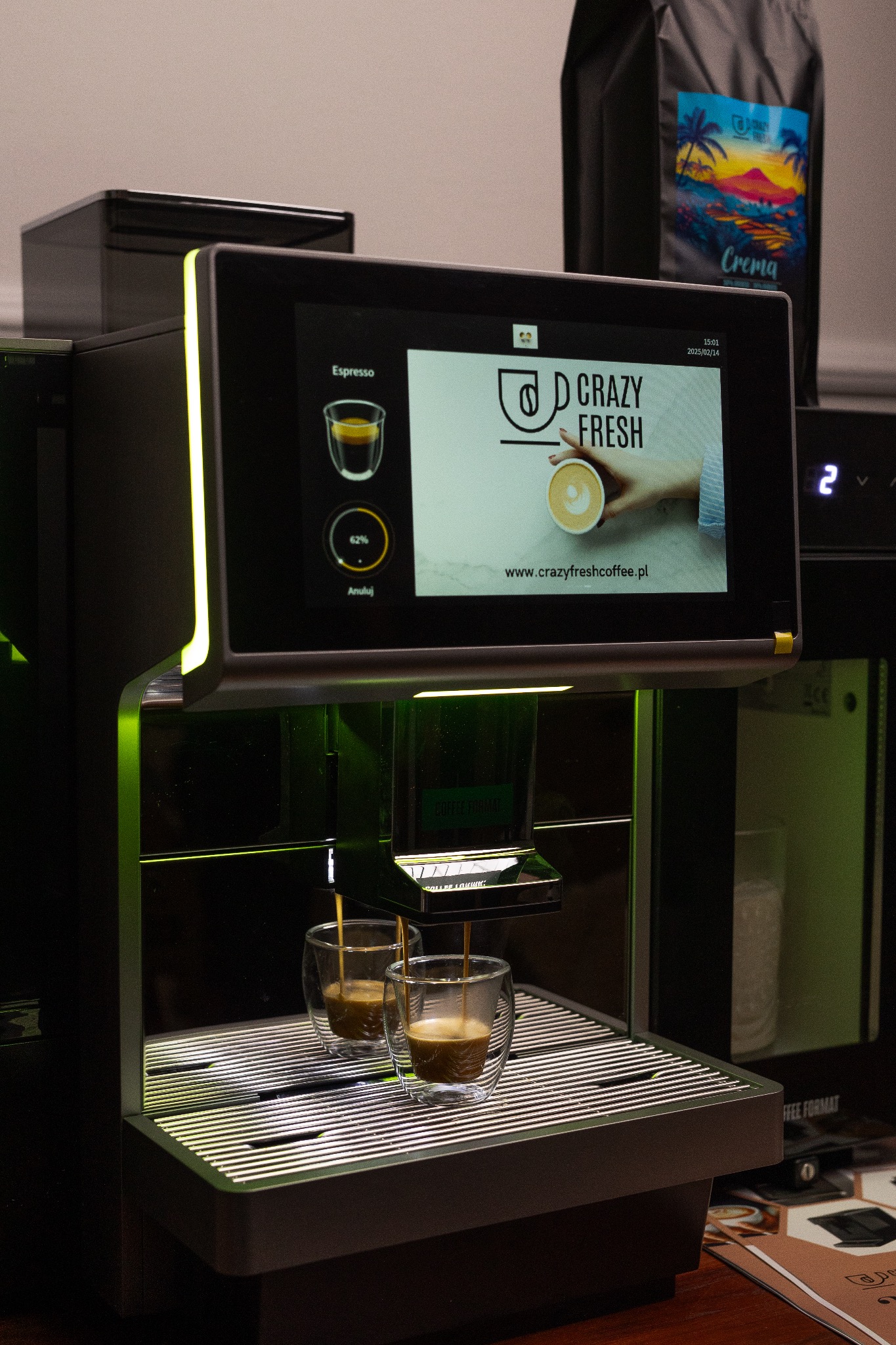 Nowoczesny ekspres do kawy z ekranem dotykowym, przygotowujący espresso do dwóch szklanek, z widocznym logiem 'Crazy Fresh' na wyświetlaczu i paczką kawy w tle.