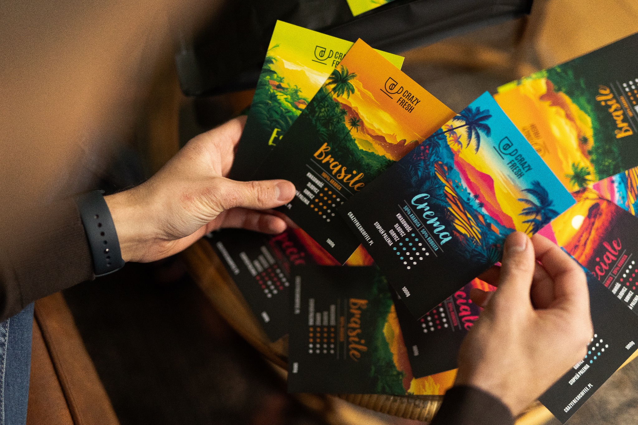 Dłonie prezentują kolekcję barwnych opakowań kawy ziarnistej marki Crazy Fresh, z motywami tropikalnych krajobrazów i informacjami o smaku, kwasowości, słodyczy i goryczy.