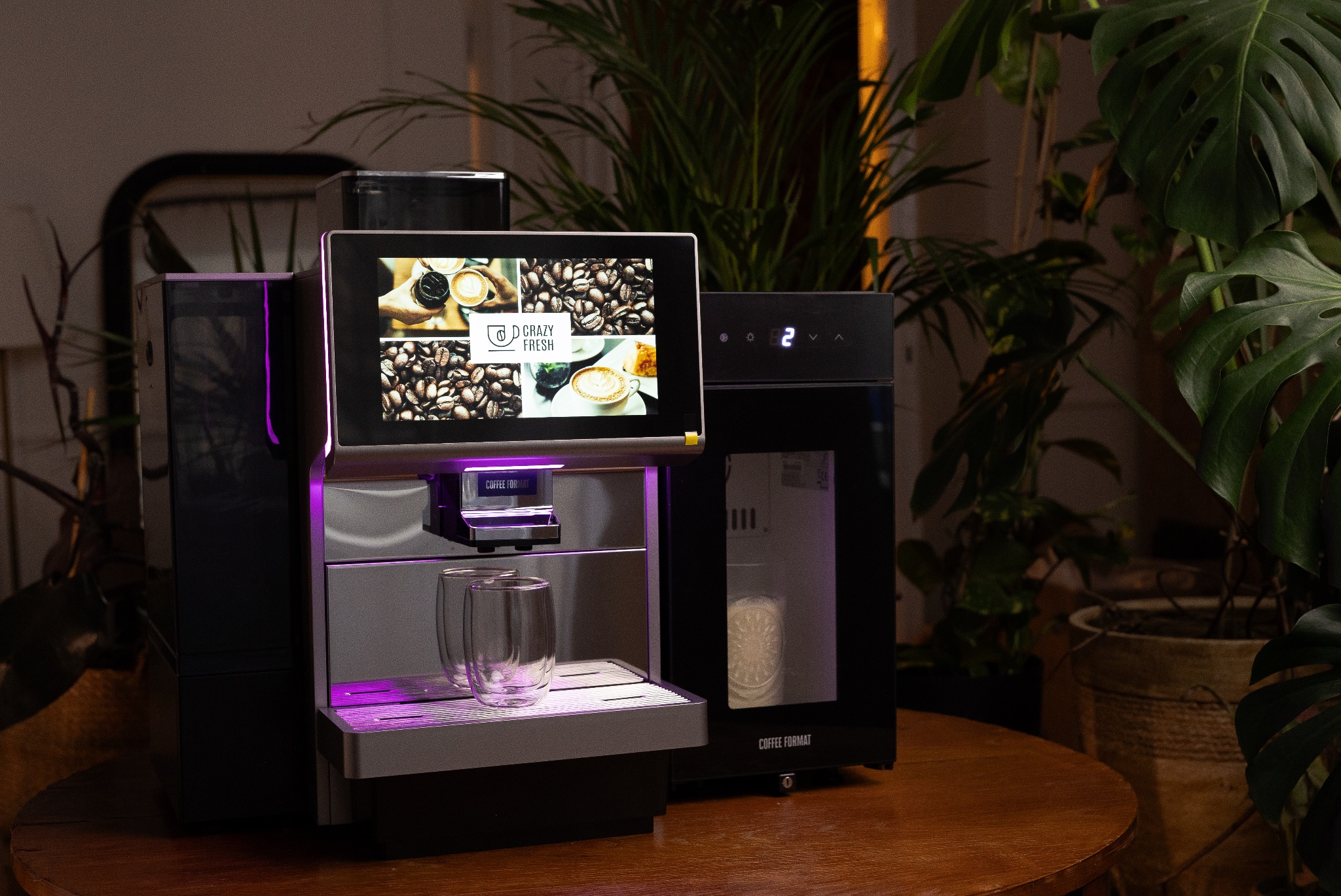 Profesjonalny ekspres do kawy z ekranem dotykowym prezentujący menu, podświetlony na fioletowo, z dwoma szklankami na podstawce, ustawiony na drewnianym stole w otoczeniu roślin doniczkowych.