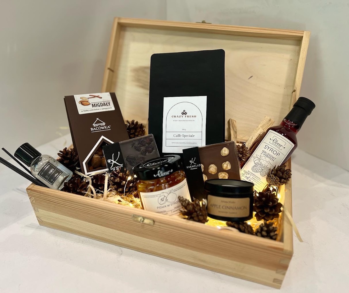 Drewniana skrzynka prezentowa wypełniona kawą, czekoladą, syropem malinowym z imbirem, konfiturą z pigwy, świecą zapachową, czekoladkami Karmello, orzechami, szyszkami i lampkami led, widok z góry.