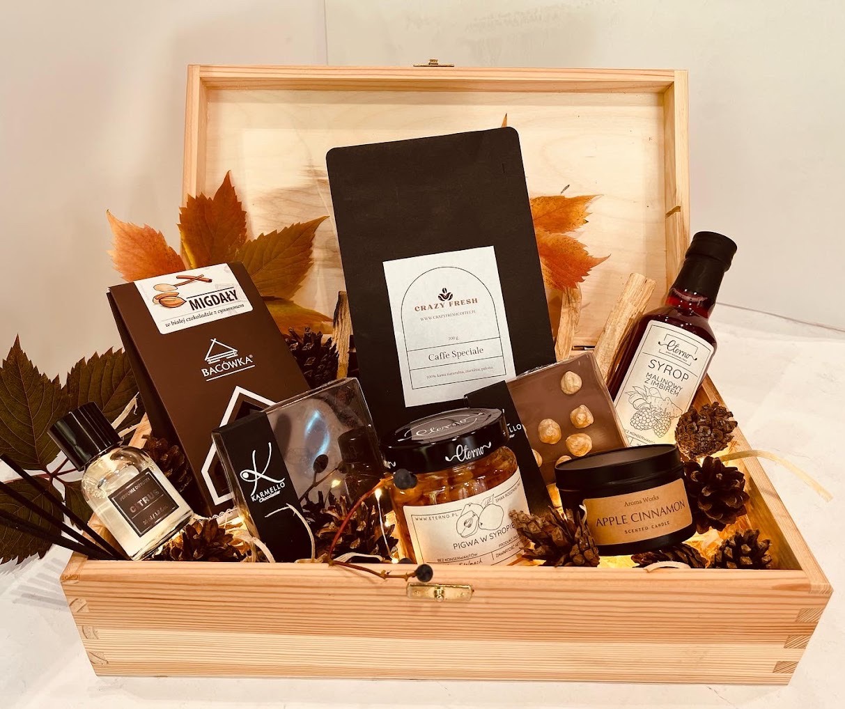 Drewniana skrzynka wypełniona upominkami: kawa, czekolada z migdałami, syrop malinowo-imbirowy, świeca zapachowa o aromacie jabłka i cynamonu, perfumy oraz dekoracyjne szyszki i jesienne liście.