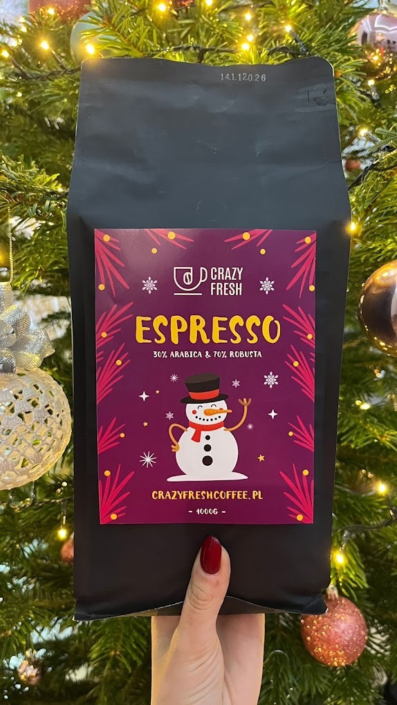 Czarna paczka kawy Espresso 30% Arabica i 70% Robusta marki Crazy Fresh z motywem świątecznym (bałwanek, śnieżynki) trzymana w dłoni na tle choinki z lampkami i bombkami.