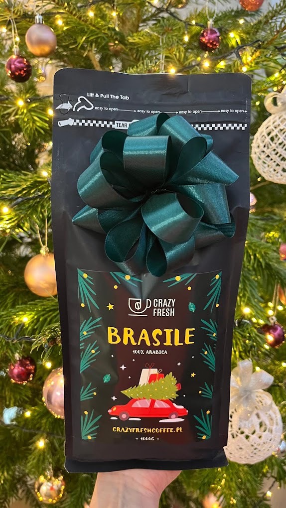 Czarna paczka kawy 'Brasile 100% Arabica' od Crazy Fresh, z zieloną kokardą, na tle choinki z ozdobami, prezentowana w dłoni.