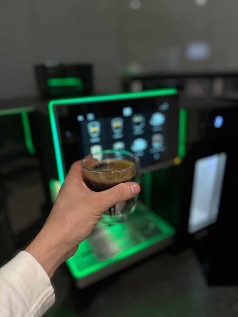 Ręka trzyma szklankę espresso przed podświetlonym na zielono ekspresem do kawy z dotykowym ekranem.