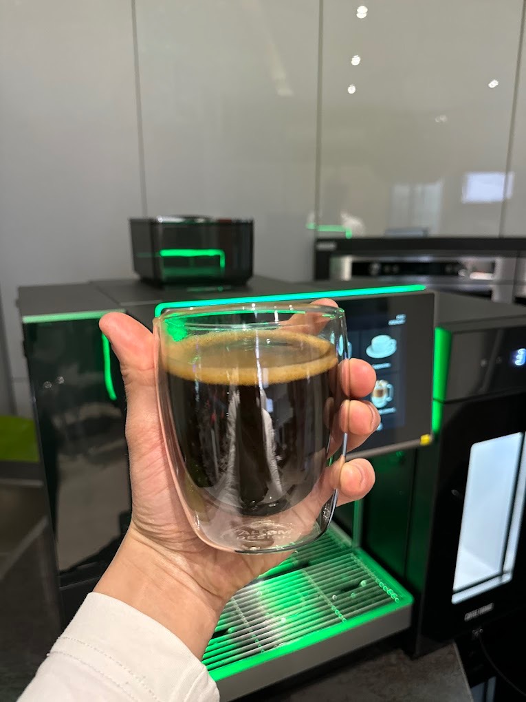 Ręka trzyma szklankę świeżo zaparzonej kawy na tle nowoczesnego ekspresu z podświetleniem LED, widoczny blat kuchenny i białe szafki.