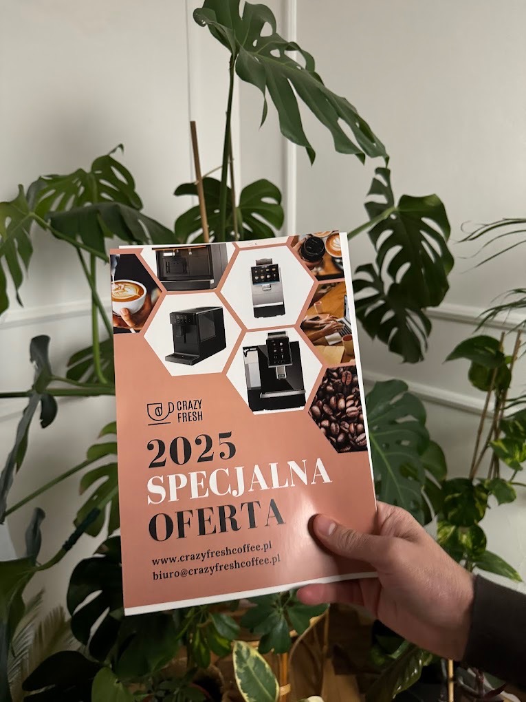 Ręka trzyma ulotkę reklamową firmy Crazy Fresh Coffee z ofertą ekspresów do kawy na rok 2025, na tle roślin doniczkowych i białej ściany.