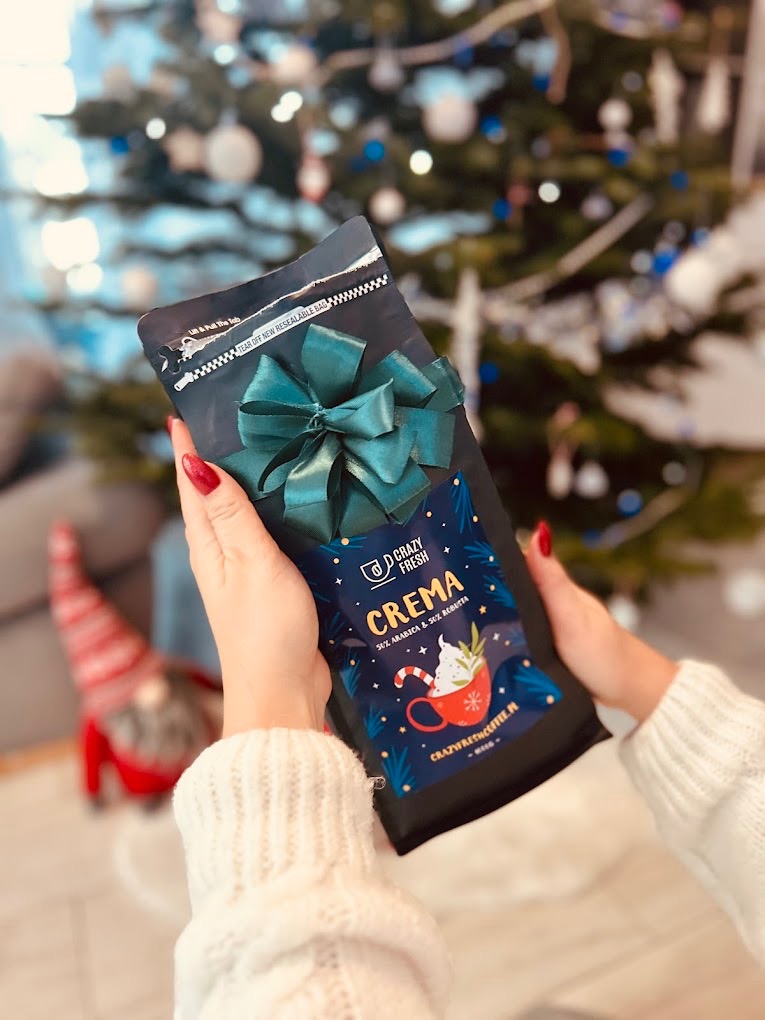 Dłonie w białym swetrze trzymają czarne opakowanie kawy 'Crazy Fresh Crema' z zieloną kokardą na tle rozświetlonej choinki i świątecznego skrzata. Opakowanie ma ilustrację świątecznej filiżanki...