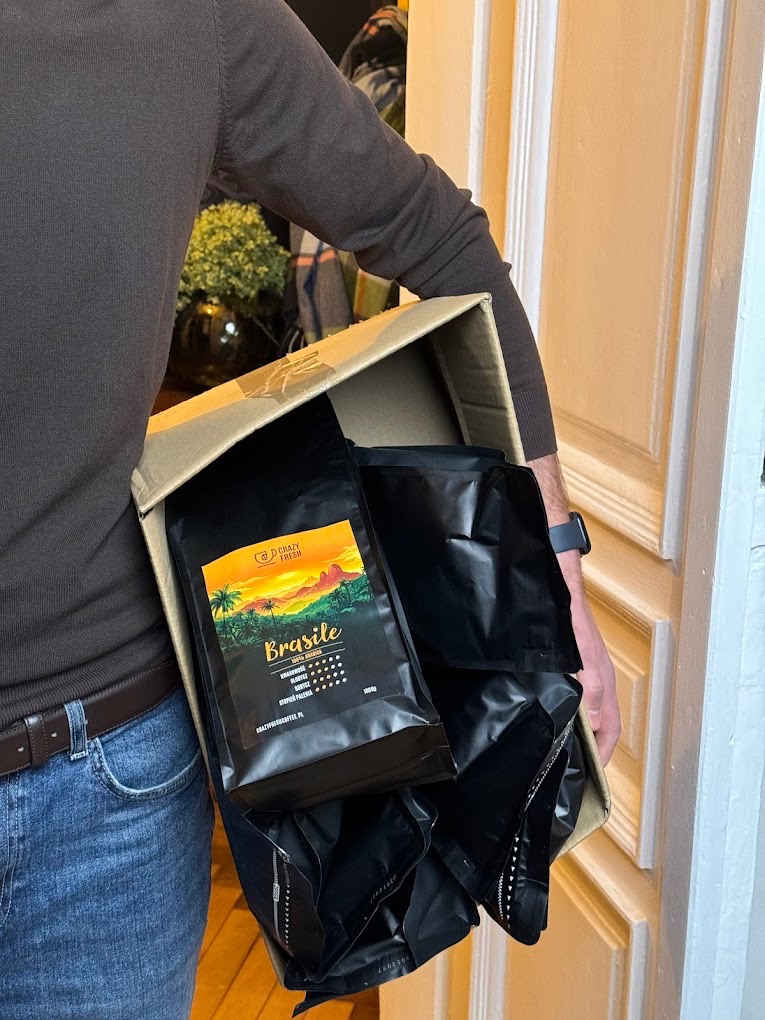 Kurier w brązowym swetrze i jeansach trzyma karton wypełniony szczelnie zapakowanymi czarnymi torbami kawy 'Brasile 100% Arabica' marki Crazy Fresh.