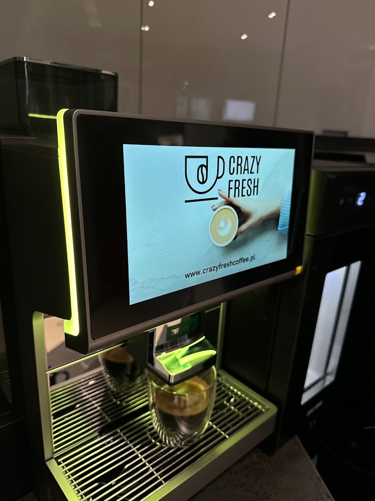 Automatyczny ekspres do kawy z podświetlanymi elementami, przygotowujący espresso w szklance z podwójnymi ściankami; na ekranie logo 'Crazy Fresh Coffee' i adres strony internetowej.