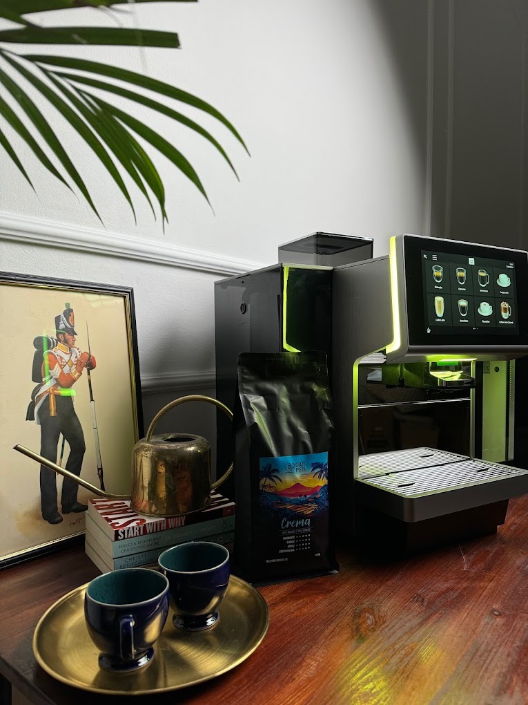 Nowoczesny ekspres do kawy z dotykowym ekranem, obok paczka kawy Crema, dwie filiżanki na złotej tacy, konewka i obrazek żołnierza na stercie książek, całość oświetlona zielonym światłem.